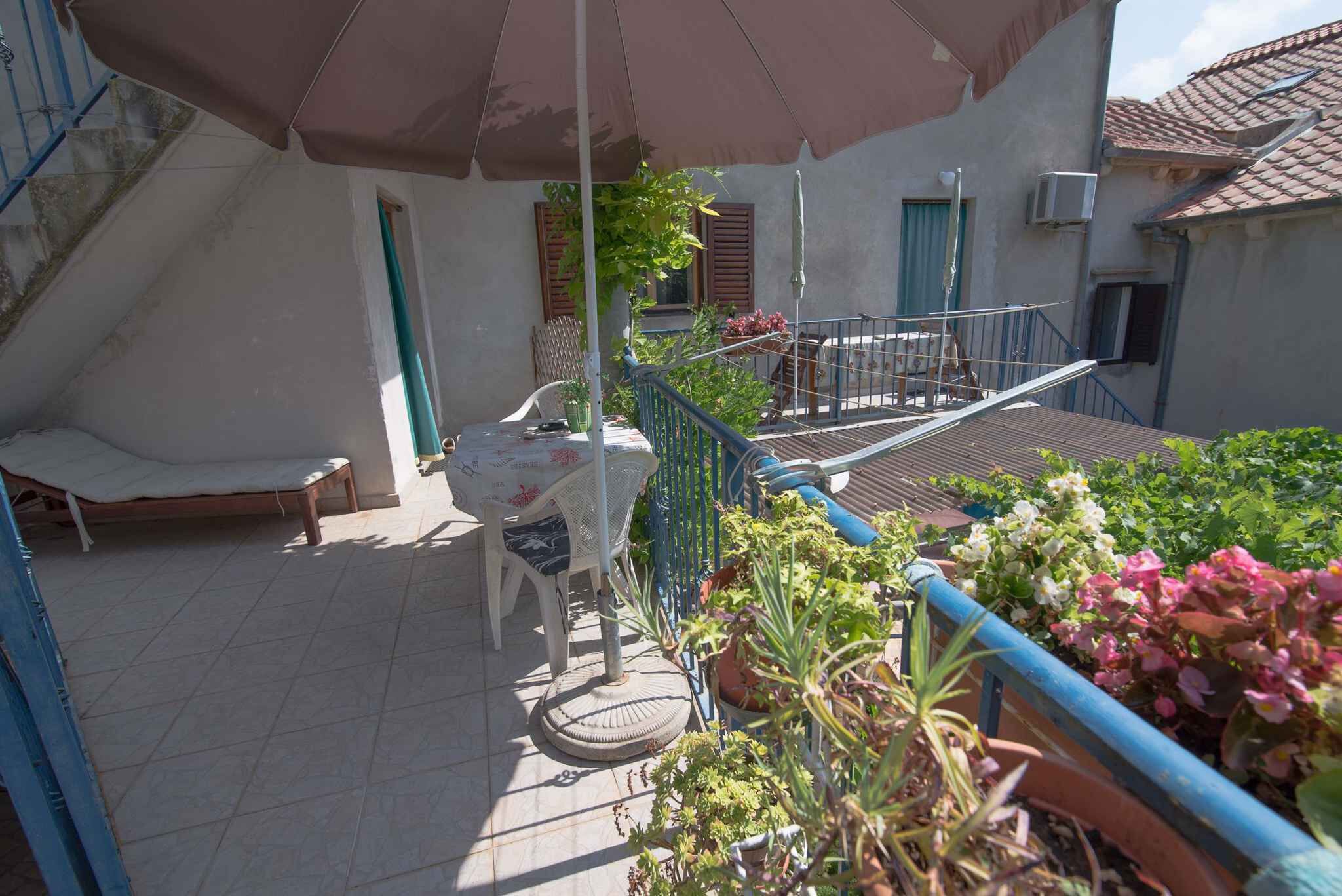 En balkon med seng, bord og stole, omgivet af krukker med planter og et blåt gelænder.