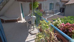 Een balkon met een bed, een tafel en stoelen, omgeven door potplanten en een blauwe balustrade.