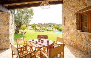 Eine Terrasse mit Steinwänden, einem runden Holztisch und Stühlen, einer hängenden Lampe sowie Blick auf Pool und Garten im Hintergrund.