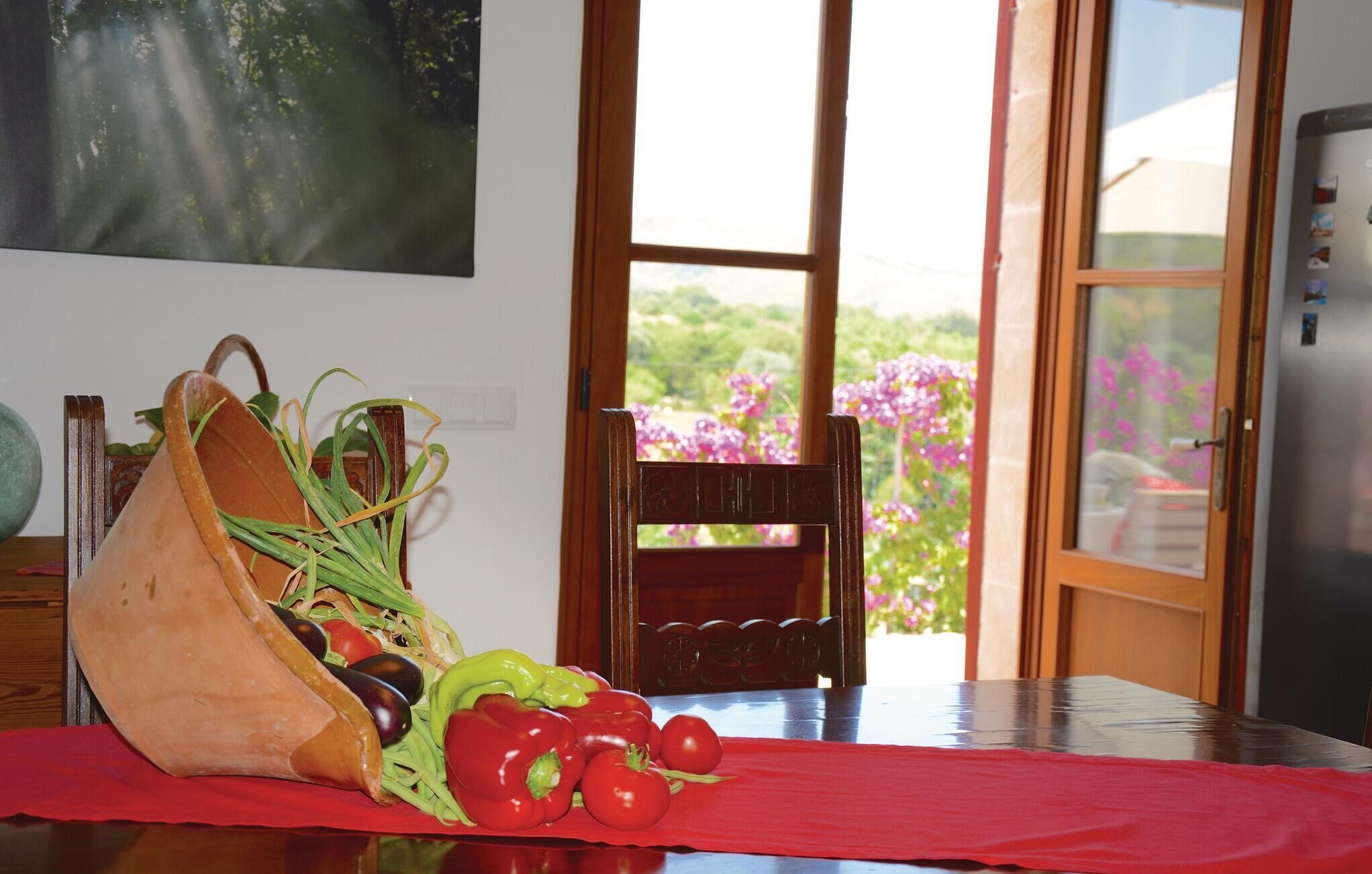 Uma mesa de jantar com toalha vermelha, um saco de legumes e uma cadeira com vista para um jardim através de uma porta de vidro.