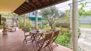Un porche cubierto con muebles de madera, plantas en macetas y vistas del jardín y edificios circundantes.