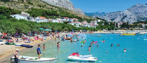 Enjoy the serene beach scene with clear blue waters, umbrellas, and sunbeds, surrounded by Mediterranean-style buildings and majestic mountains.