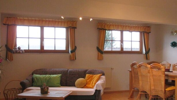 Ein Wohnzimmer mit einer Couch, einem Couchtisch, einem Esstisch mit Stühlen und zwei Fenstern mit Vorhängen.