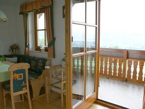 A room with a dining table, chairs, a window with curtains, and a balcony with a railing. - Apartment 2 - Ferienhof Nußbaumer (Gaisberg)