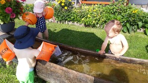Kinder spielen in der Nähe einer hölzernen Wasserspielsatzes, im Hintergrund sind ein Tisch und Stühle zu sehen.