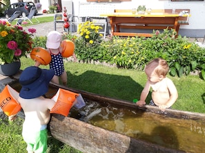 Des enfants jouent près d’une fontaine en bois, avec une table et des chaises en arrière-plan.