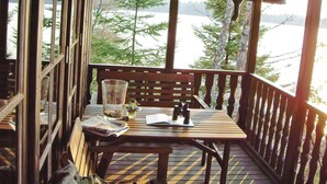 Eine Holzterrasse mit Tisch, Stuhl und Bank. Ein Fenster bietet Blick auf Bäume und ein Gewässer.