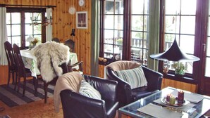 Ein gemütlicher Raum mit Holzwänden, einem Essbereich mit Tisch und Stühlen, einem Sofa mit gestreiftem Kissen und einem großen Fenster mit Vorhängen.