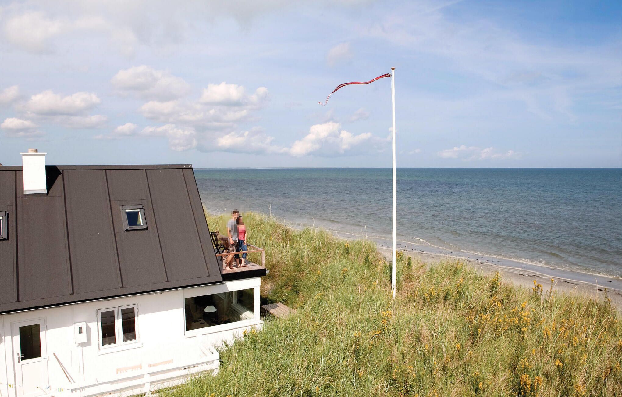Una casa costera con un balcón que da al mar, un asta con una bandera y una zona verde al frente.