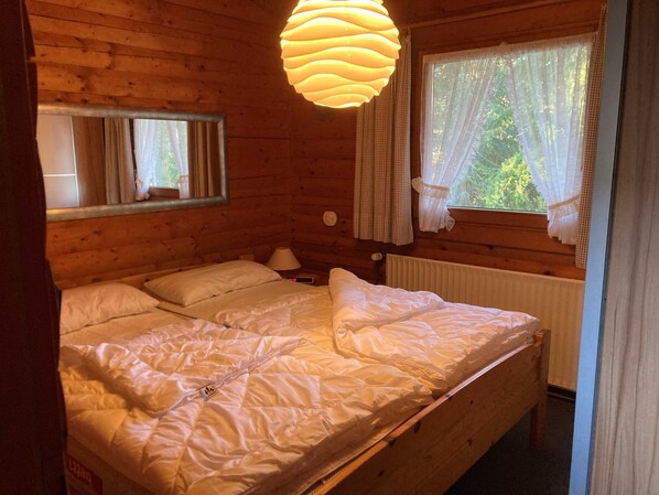 A wooden cabin bedroom with a bed, a nightstand, a lamp, a window with curtains, and a mirror.
