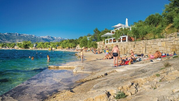 Una playa con gente tomando el sol y nadando, un muro de piedra y un edificio con techo blanco al fondo.
