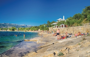 En strand med folk som solar och badar, en stenmur och en byggnad med ett vitt tak i bakgrunden.