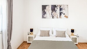 Un dormitorio con una cama, dos mesitas de noche, una obra de arte en la pared y un ventanal con cortinas.