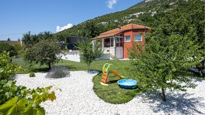 A house with a red exterior and a balcony, surrounded by a garden with a play structure and a blue pool.