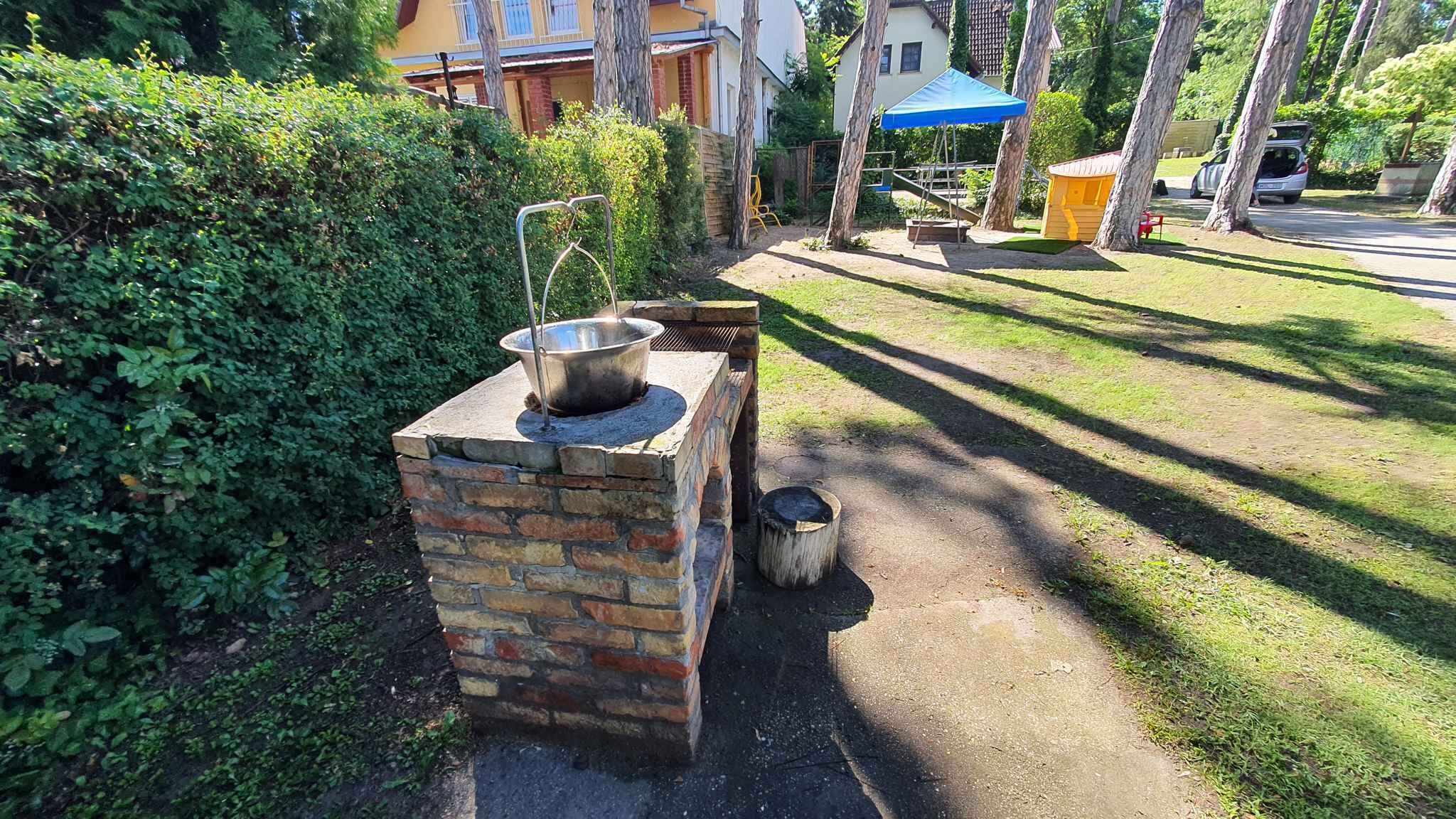 A brick outdoor cooking area with a metal pot on a stove, surrounded by greenery and a paved path.