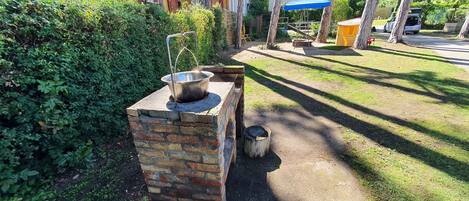 A brick outdoor cooking area with a metal pot on a stove, surrounded by greenery and a paved path.