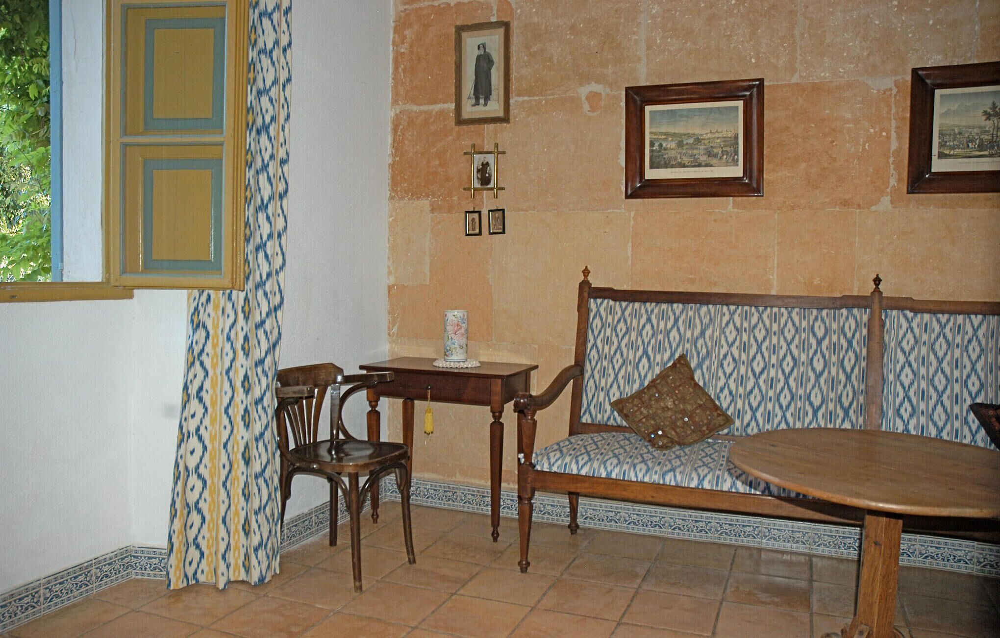 A room with a patterned sofa, a wooden table, and a chair. There are framed pictures on the wall and a window with a patterned curtain.