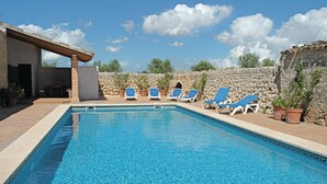 A swimming pool with blue water, surrounded by stone walls and potted plants. There are several blue lounge chairs along the poolside.