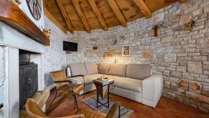 A cozy living room with a stone wall, wooden ceiling beams, a sofa, two leather chairs, a small table with fruit, and a television mounted on the wall. - Lovely home in Cukrici with WiFi (Cukrici)