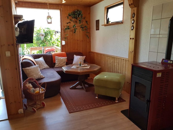 A cozy cabin interior with a wooden floor, a sofa, a small table, a green ottoman, and a wood-burning stove.