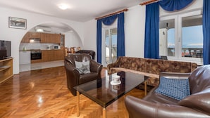 Un salon avec un canapé, deux fauteuils, une table basse en verre et une cuisine en arrière-plan.