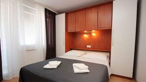 A hotel room with a bed, wooden headboard, white curtains, and a window with blinds. - Holiday apartment near the sea (Baška)