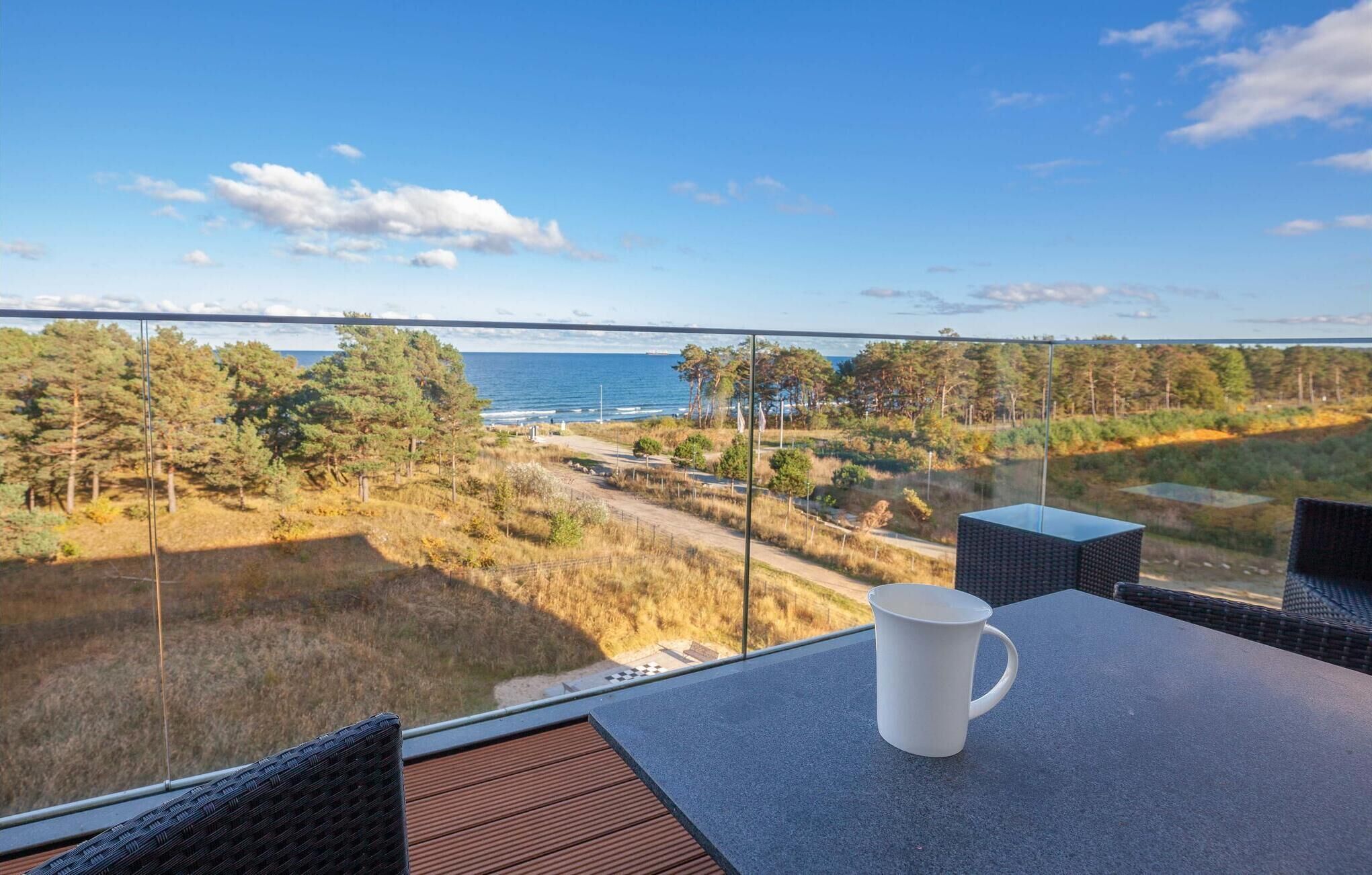 A balcony with a table, a white mug, and a view of the sea and forest.