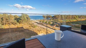 En balkong med ett bord, en vit mugg och utsikt över havet och skogen.