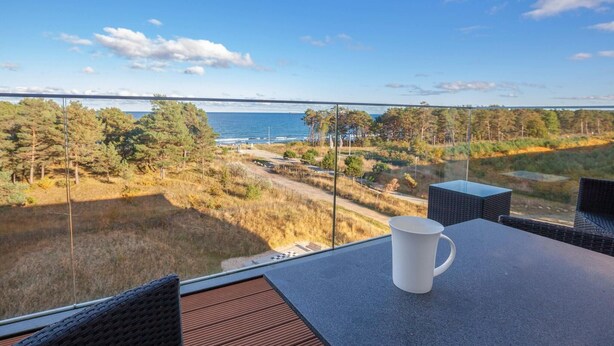 A balcony with a table, a white mug, and a view of the sea and forest. - Beautiful apartment in Binz with outdoor swimming pool (Prora/Rügen)