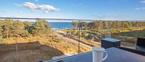 A balcony with a table, a white mug, and a view of the sea and forest.