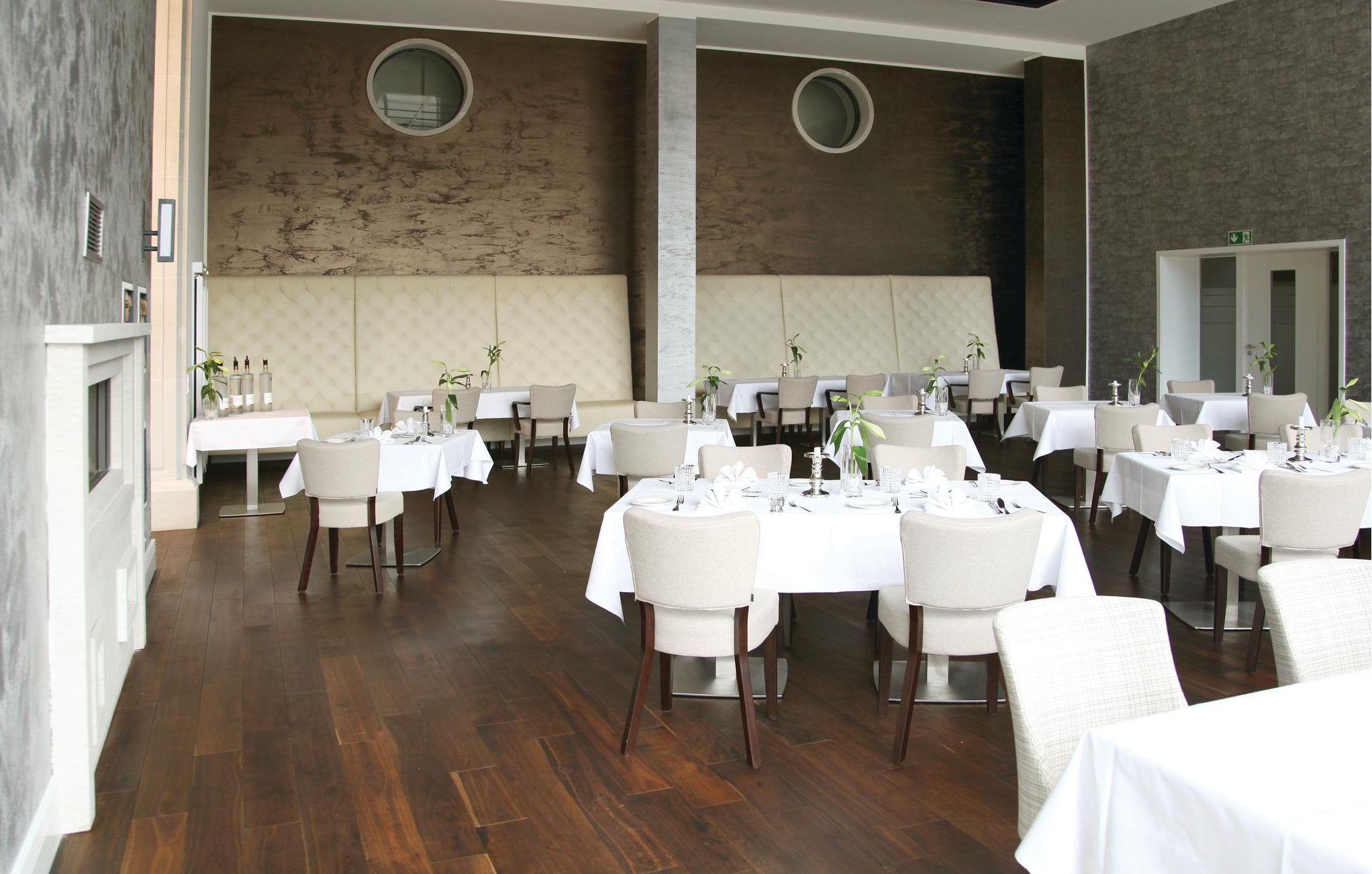 A restaurant with round tables and chairs, white tablecloths, and a wooden floor.