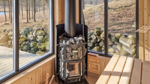 Ein Kaminofen in einer Hütte mit großen Fenstern, die Blick auf eine Steinwand und Bäume gewähren.