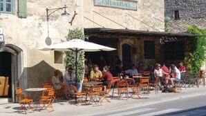 Un café al aire libre con mesas y sillas, un cartel sobre la entrada y un edificio rústico de piedra al fondo.