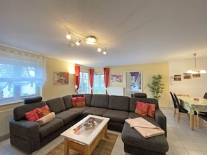 Uma sala de estar com um sofá seccional, uma mesa de centro de vidro e um espaço de jantar com mesa e cadeiras.