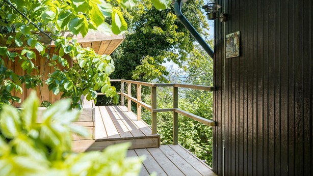 Eine Holzterrasse mit Metallgeländer, die zu einem dunklen Holzgebäude mit einem kleinen Fenster führt.
