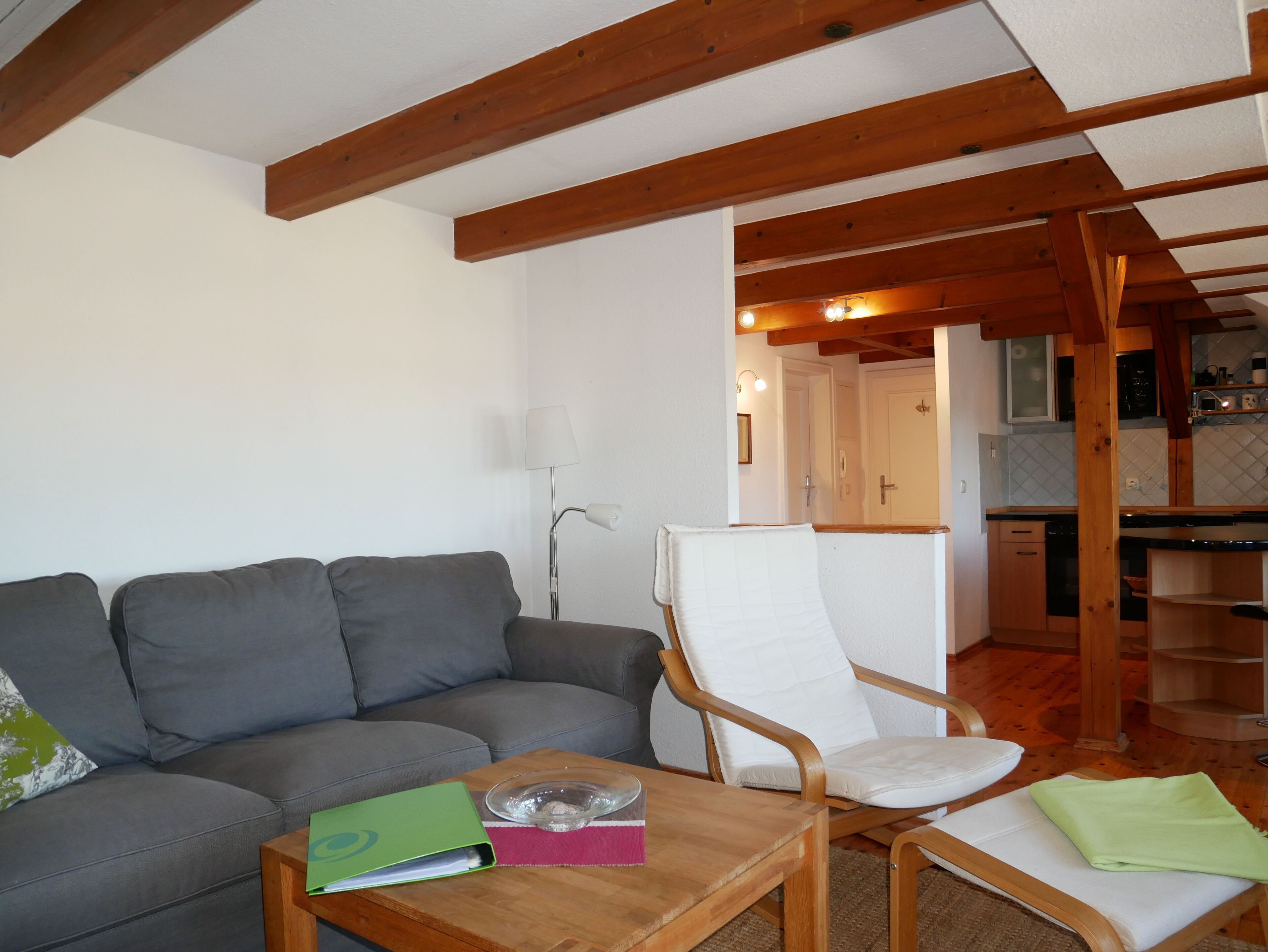 A living room with a grey sofa, a wooden coffee table, and a white armchair. There is a kitchen visible in the background.