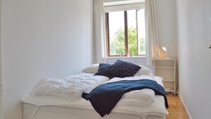 A small, neatly made bed with white and dark blue bedding, a bedside table with a lamp, and a window with curtains. - 1 bedroom stunning apartment in Hornbæk (Hornbæk)