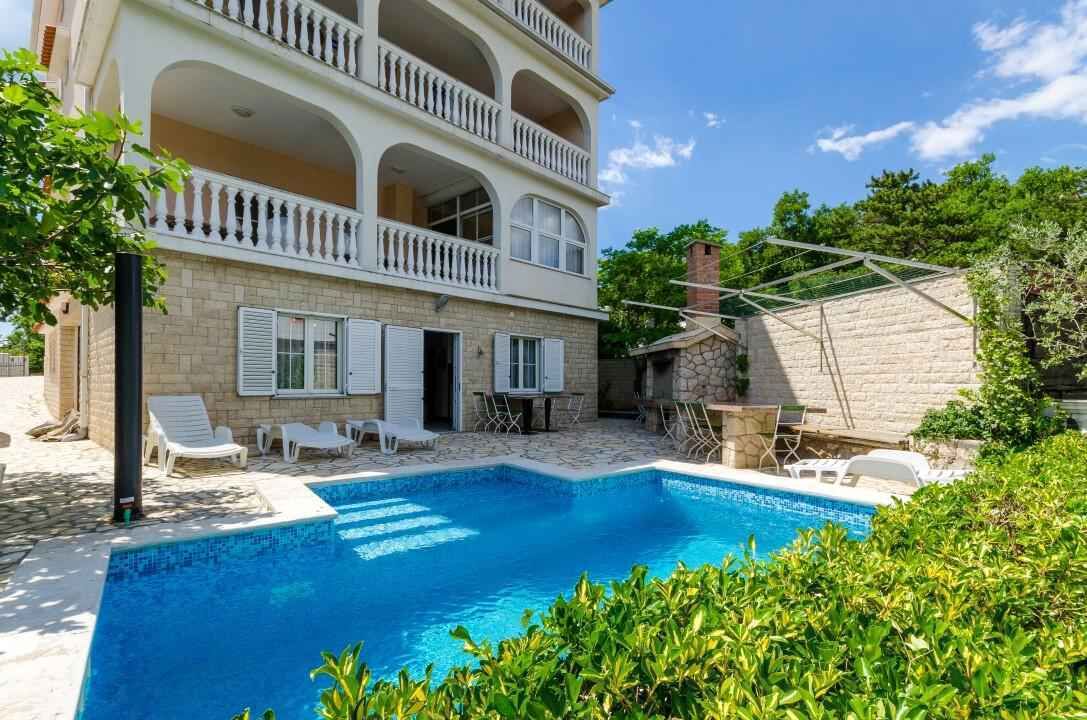 A multi-story building with a swimming pool, outdoor seating, and a stone wall.