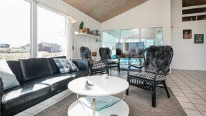 A modern living room with a black leather sofa, two black wicker chairs, a white coffee table, and a glass-top dining table. Large windows offer a view of the beach and houses. - Amazing home in Løkken with sauna (Løkken)