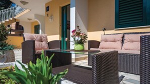 Un área de estar al aire libre con mobiliario de mimbre y una mesa de vidrio, rodeada de plantas en macetas y una escalera.