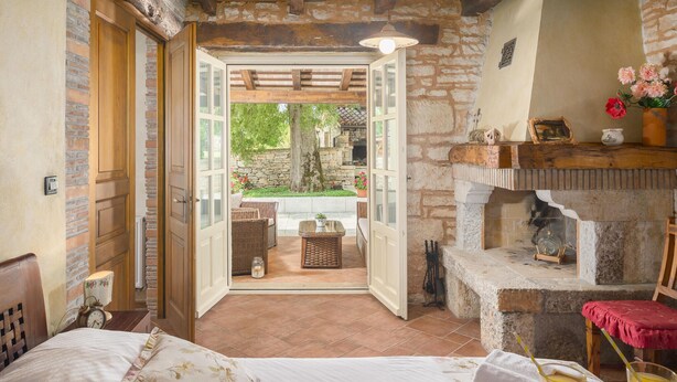 A cozy room with a bed, a breakfast tray, a stone fireplace, and an open door leading to an outdoor area with wicker furniture and a garden. - Stunning home in Tinjan with sauna (Tinjan)