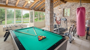 A pool table with billiard balls, a treadmill, and a punching bag in a room with stone walls and large windows. - Stunning home in Tinjan with sauna (Tinjan)