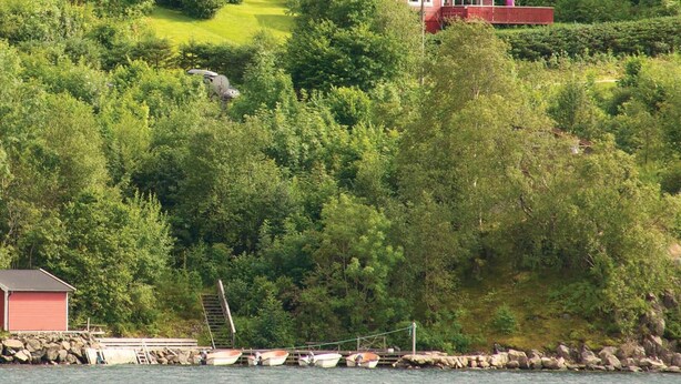 Une cabane rouge dotée d’un balcon, entourée d’une végétation luxuriante et bordée d’un quai où sont amarrés des bateaux.