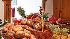Un tavolo da buffet con diversi tipi di pane, frutta e un contenitore di plastica trasparente pieno di cibo.