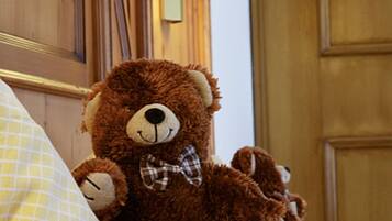 A brown teddy bear sitting on a bed with a white pillow and a lamp in the background.