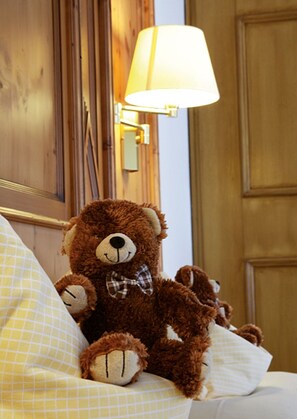 Um urso de pel úcia marrom sentado em uma cama com um travesseiro branco e uma luminária ao fundo.