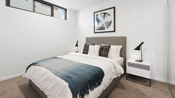 A modern bedroom with a large bed, a nightstand, a framed picture on the wall, and a window with blinds.