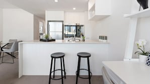 A modern kitchen with white cabinetry, a central island with stools, and a view of the cityscape through the window. - Ivy and Eve Apartments by CLLIX (South Brisbane)