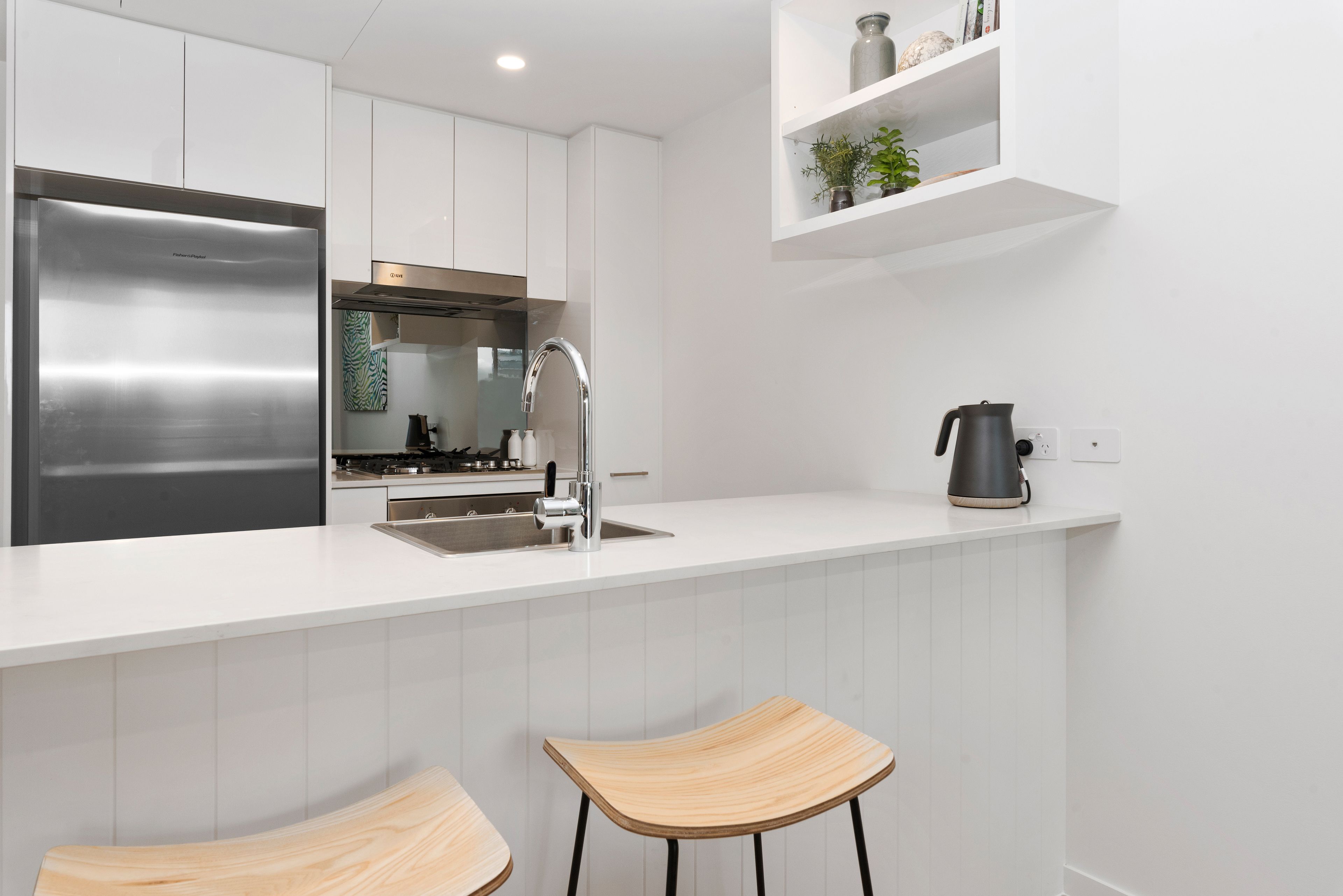 A modern kitchen with white cabinetry, a stainless steel refrigerator, a gas stove, and two wooden bar stools.