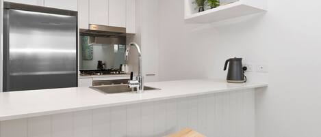 Una cocina moderna con armarios blancos, un refrigerador de acero inoxidable, una estufa a gas y dos taburetes de madera para bar.
